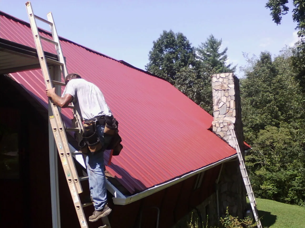Roofer on a ladder beside a red metal roof and stone chimney — Soffit & Fascia Repair by Roofing Brothers in East Windsor